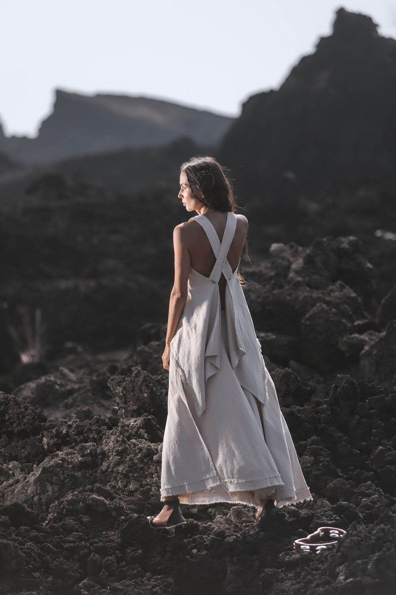 A woman wearing the AYA Sacred Wear Off-White Linen Boho Top for Women • Viking Tunic stands on rocky terrain, gazing into the distance; the dark, rugged landscape features blurred mountains in the background.