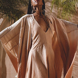 In a sunny outdoor setting, a woman in a Powder Pink Boho Poncho by AYA Sacred Wear stands with arms outstretched and eyes closed, facing upward. Palm fronds and a stone wall create a tropical vibe in the background.