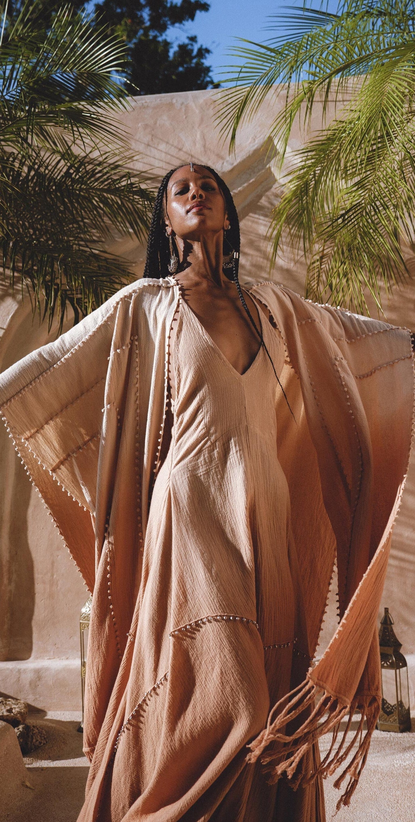 In a sunny outdoor setting, a woman in a Powder Pink Boho Poncho by AYA Sacred Wear stands with arms outstretched and eyes closed, facing upward. Palm fronds and a stone wall create a tropical vibe in the background.