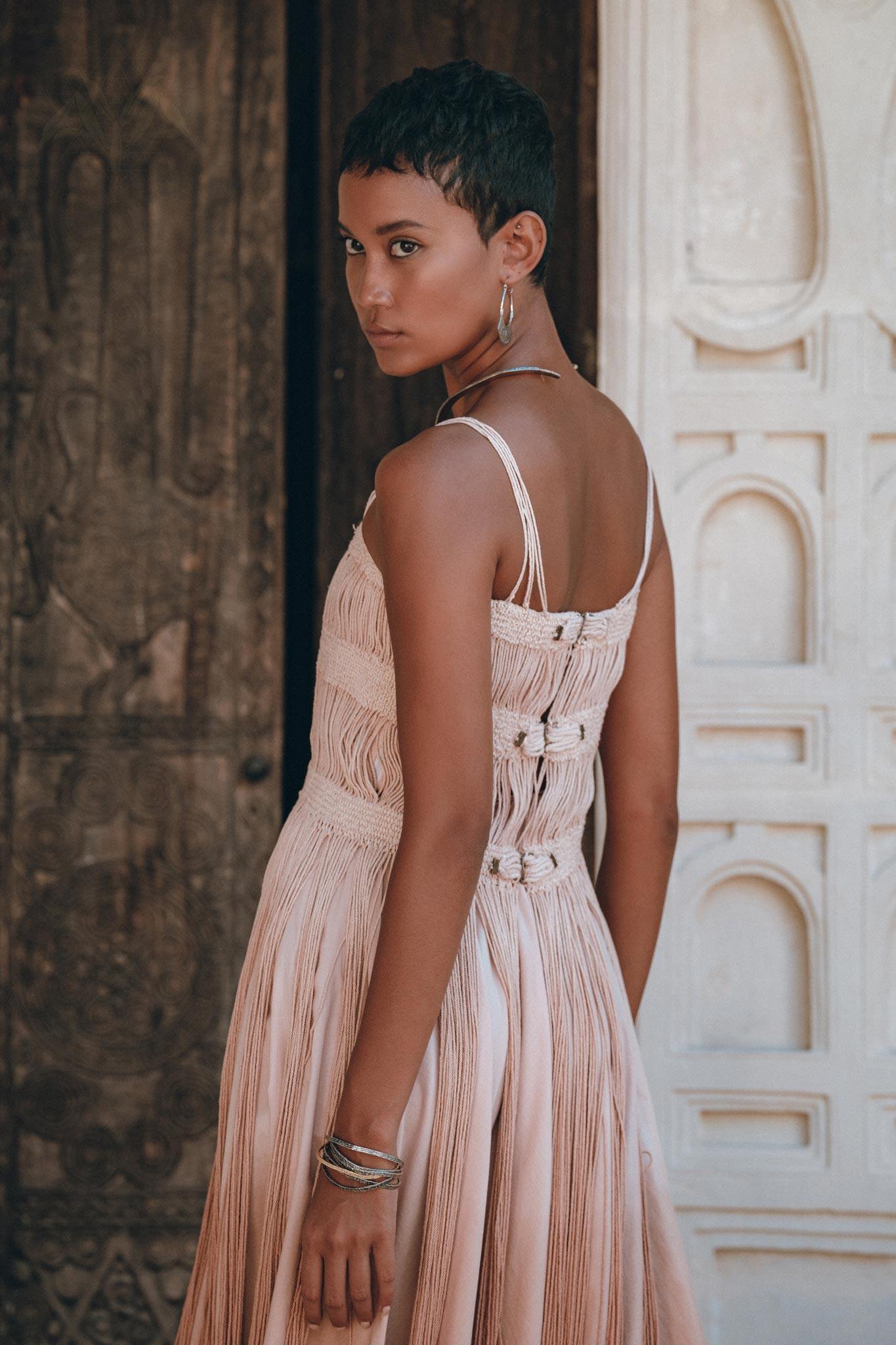 A person with short hair wears the AYA Sacred Wear Dusty Pink Tribal Goddess Slip Over net overlay dress., an ethnic handwoven macramé sheer net layer. Posed sideways with hoop earrings and bracelets, they stand before ornate wooden and white carved doors.