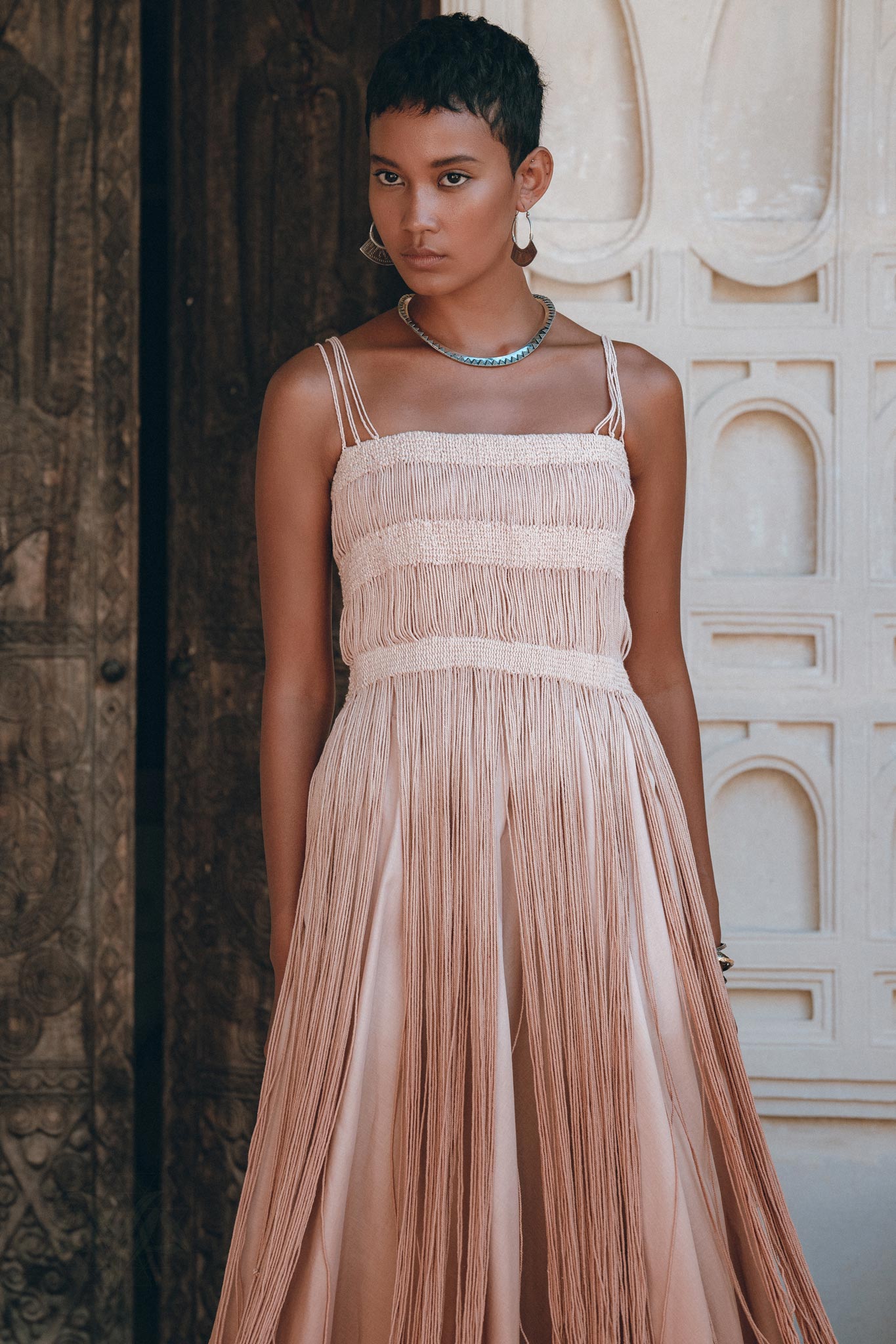 A woman with short hair wears the AYA Sacred Wear Dusty Pink Tribal Goddess Slip Over net overlay dress, an ethnic handwoven macrame layer-up sheer net dress, accessorized with hoop earrings and a beaded necklace, standing before an ornate door.