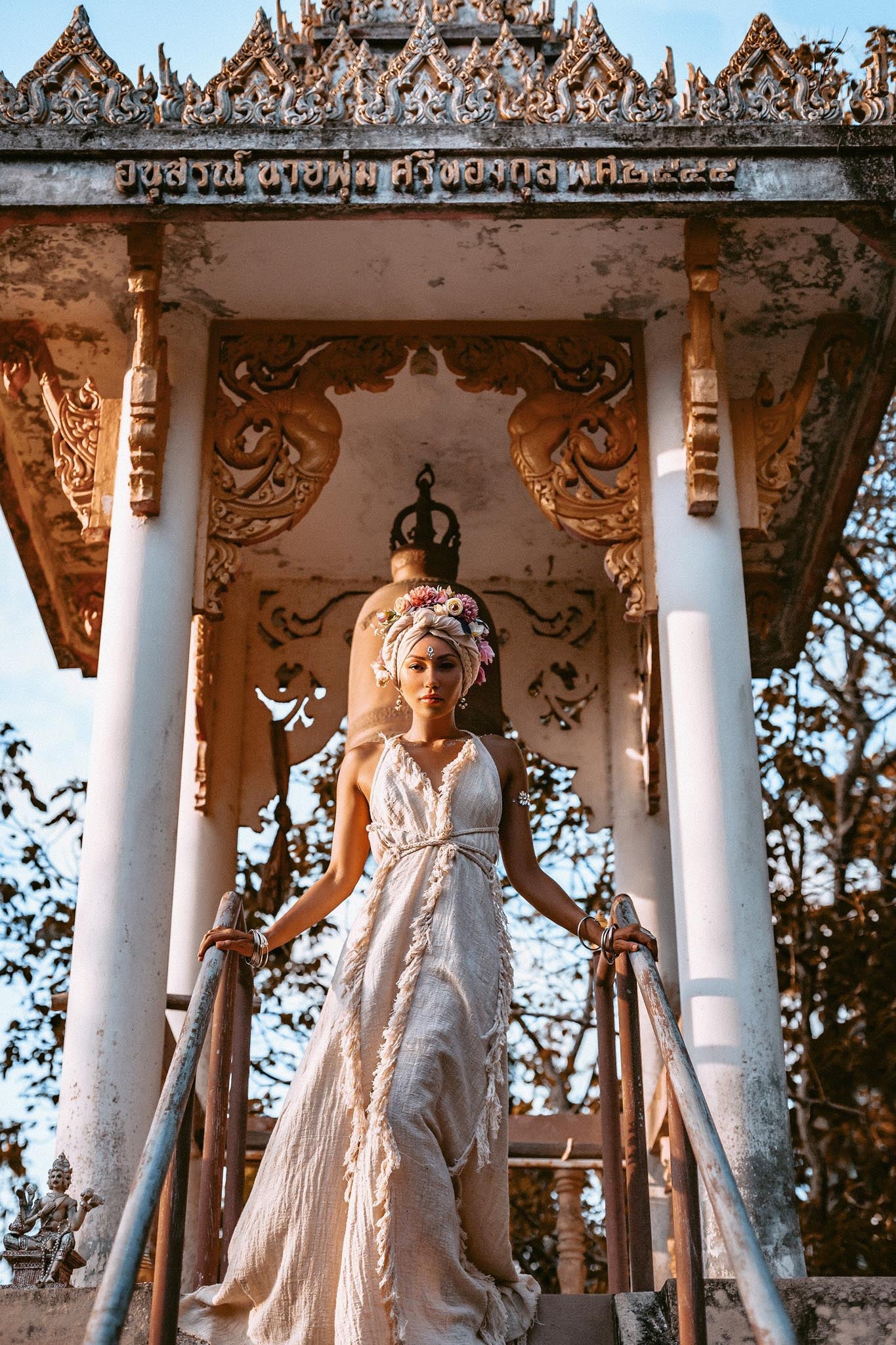 A bohemian bride wearing the Off-White raw cotton goddess wedding dress by AYA Sacred Wear, standing gracefully on ancient temple stairs with a floral headpiece.
