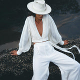 A person wearing an off-white outfit from AYA Sacred Wear, featuring the Off White Adjustable Pants – Casual Wide Boot Trousers as part of a v-neck long sleeve top with billowing sleeves, and high-waisted wrap-around pants made of an organic cotton blend, complemented by a white hat. They are seated on a rock near the water with a partially downward gaze, surrounded by a serene atmosphere.