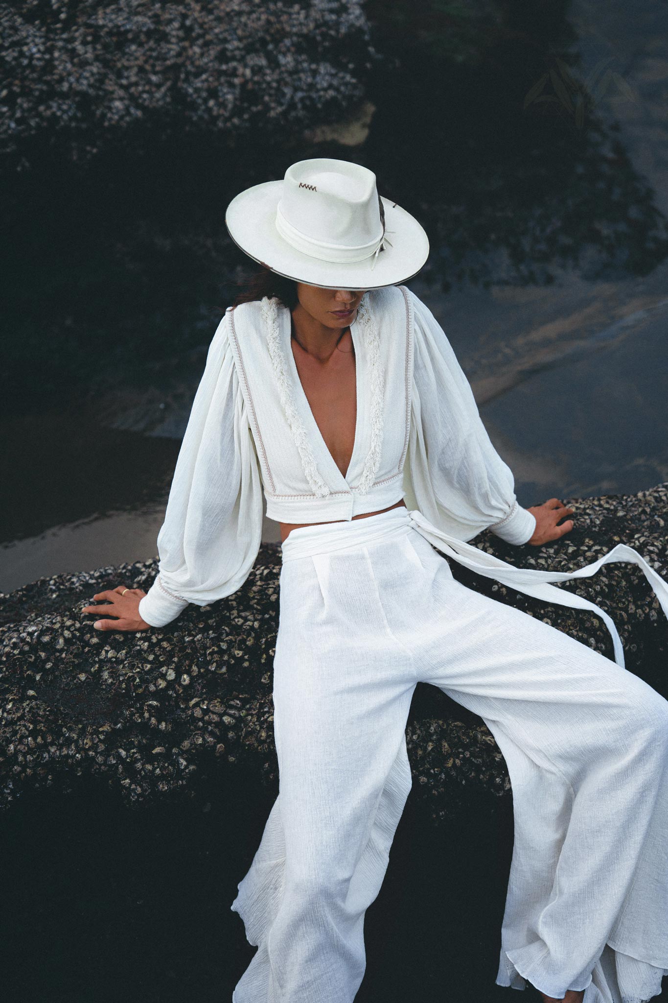 A person wearing an off-white outfit from AYA Sacred Wear, featuring the Off White Adjustable Pants – Casual Wide Boot Trousers as part of a v-neck long sleeve top with billowing sleeves, and high-waisted wrap-around pants made of an organic cotton blend, complemented by a white hat. They are seated on a rock near the water with a partially downward gaze, surrounded by a serene atmosphere.