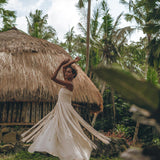 A woman in an Off-White Tribal Goddess Slip Over by AYA Sacred Wear dances joyfully outdoors near a thatched-roof hut, surrounded by lush palm trees and tropical greenery. The ethnic handwoven macrame layer adds a unique, sheer touch.
