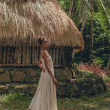 A woman wears the AYA Sacred Wear Off-White Tribal Goddess Slip Over, an ethnic handwoven macrame sheer net, standing before a thatched-roof hut amid lush greenery and palm trees.