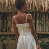 A woman in the Off-White Tribal Goddess Slip Over by AYA Sacred Wear stands outdoors, her back turned amid rustic wooden fencing, stones, and thatched roofing—a serene setting for this ethnic handwoven macrame layer up sheer net dress.