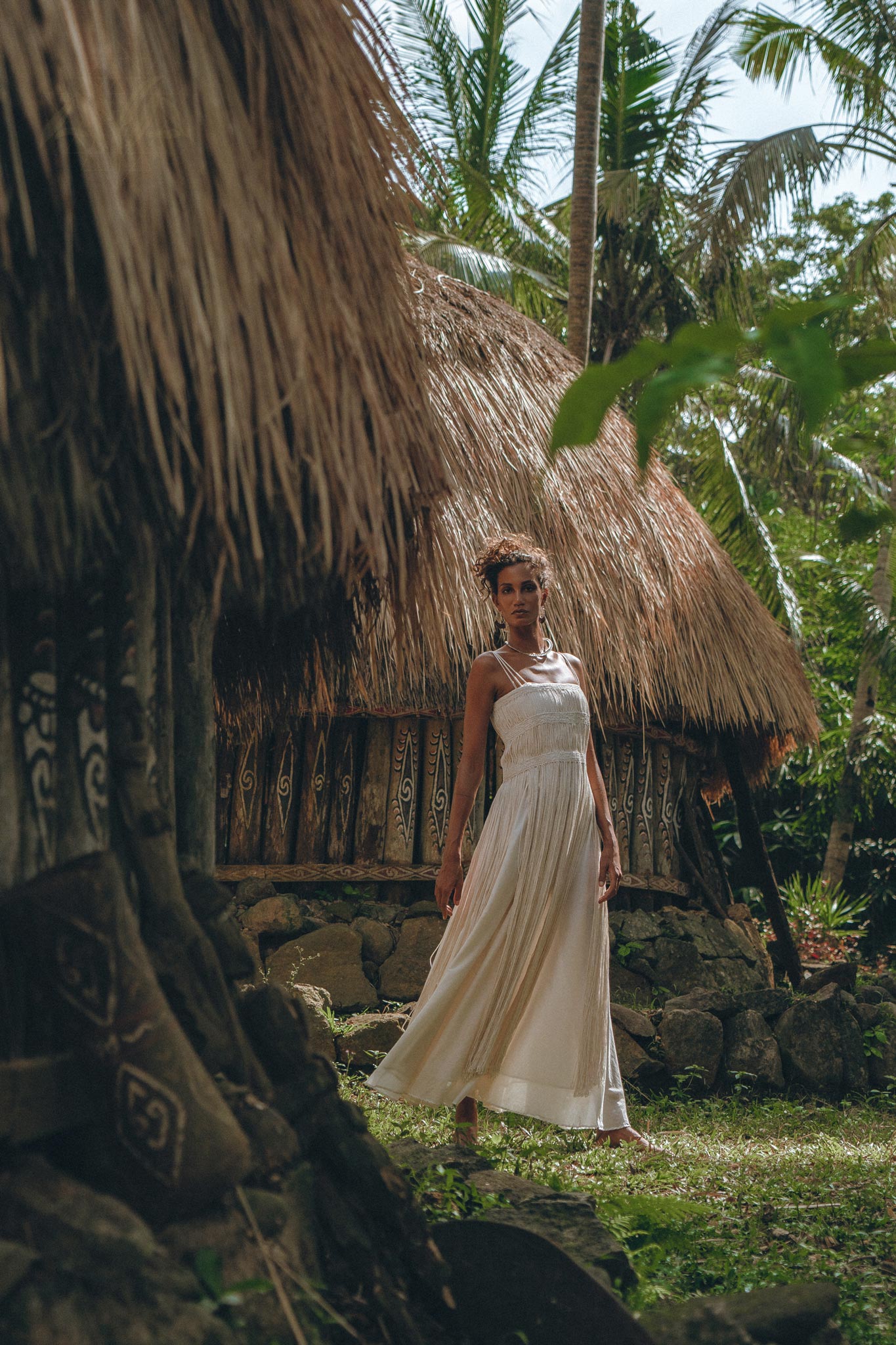 A woman in an Off-White Tribal Goddess Slip Over by AYA Sacred Wear stands on grass near a thatched-roof hut, surrounded by lush tropical plants and tall palms, wearing an ethnic handwoven macrame sheer net dress.