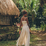A woman in the Off-White Tribal Goddess Slip Over by AYA Sacred Wear strolls barefoot near a thatched-roof hut, encircled by lush tropical greenery, showcasing the ethnic handwoven macrame and sheer net design.
