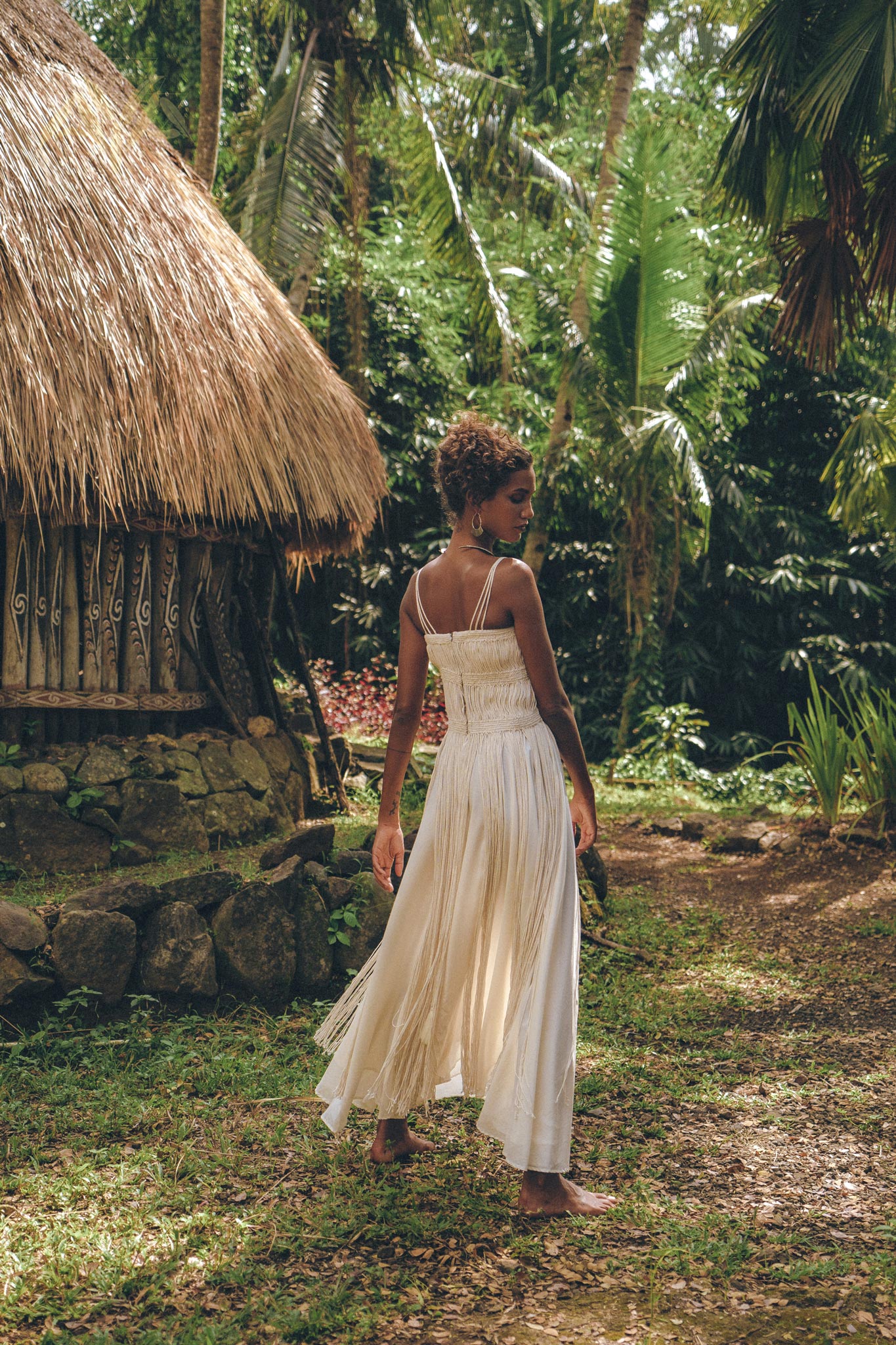 A woman in the Off-White Tribal Goddess Slip Over by AYA Sacred Wear strolls barefoot near a thatched-roof hut, encircled by lush tropical greenery, showcasing the ethnic handwoven macrame and sheer net design.