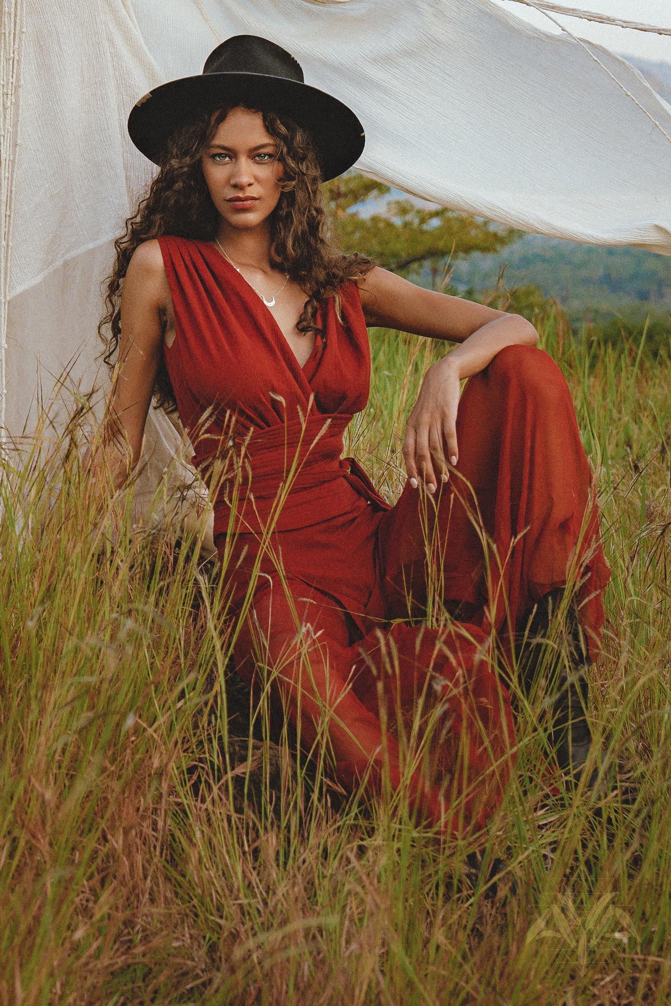 A bohemian model in a black wide-brimmed hat sits gracefully in a golden field, showcasing the flowy drape and high side slit of the Red Rose jumpsuit by AYA Sacred Wear