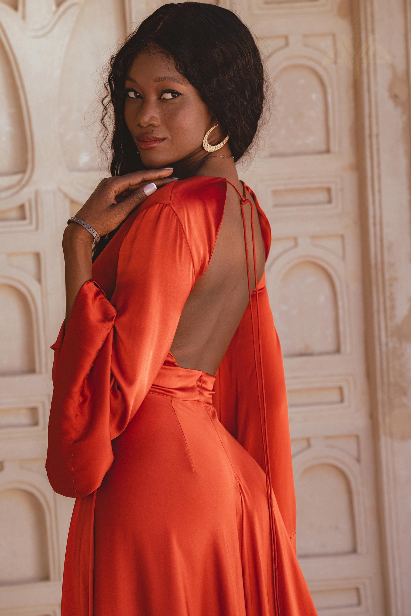 A woman poses gracefully in a wine red maxi dress with an open back, featuring long sleeves and a flowing silhouette. She is standing against a textured beige backdrop, showcasing the elegant design of the dress, perfect for a beach wedding. The look is completed with statement earrings and a stylish accessory on her wrist.