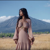 A woman with long dark hair stands in a golden field, wearing the AYA Sacred Wear Samudra Dress in Blush Rose—a backless boho maxi dress. She faces left, with distant mountains, trees, and a blue sky with scattered clouds behind her.
