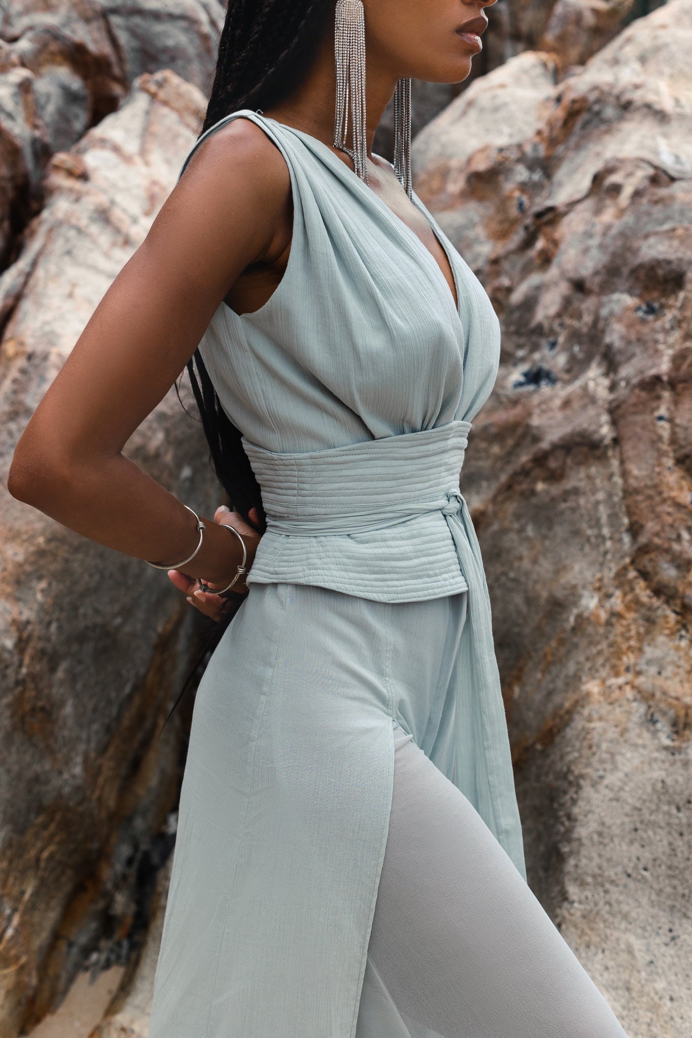 Close-up profile of a model sitting by coastal rocks, highlighting the deep V-neckline and structural belted corset of the Powder Blue organic cotton jumpsuit by AYA Sacred Wear.