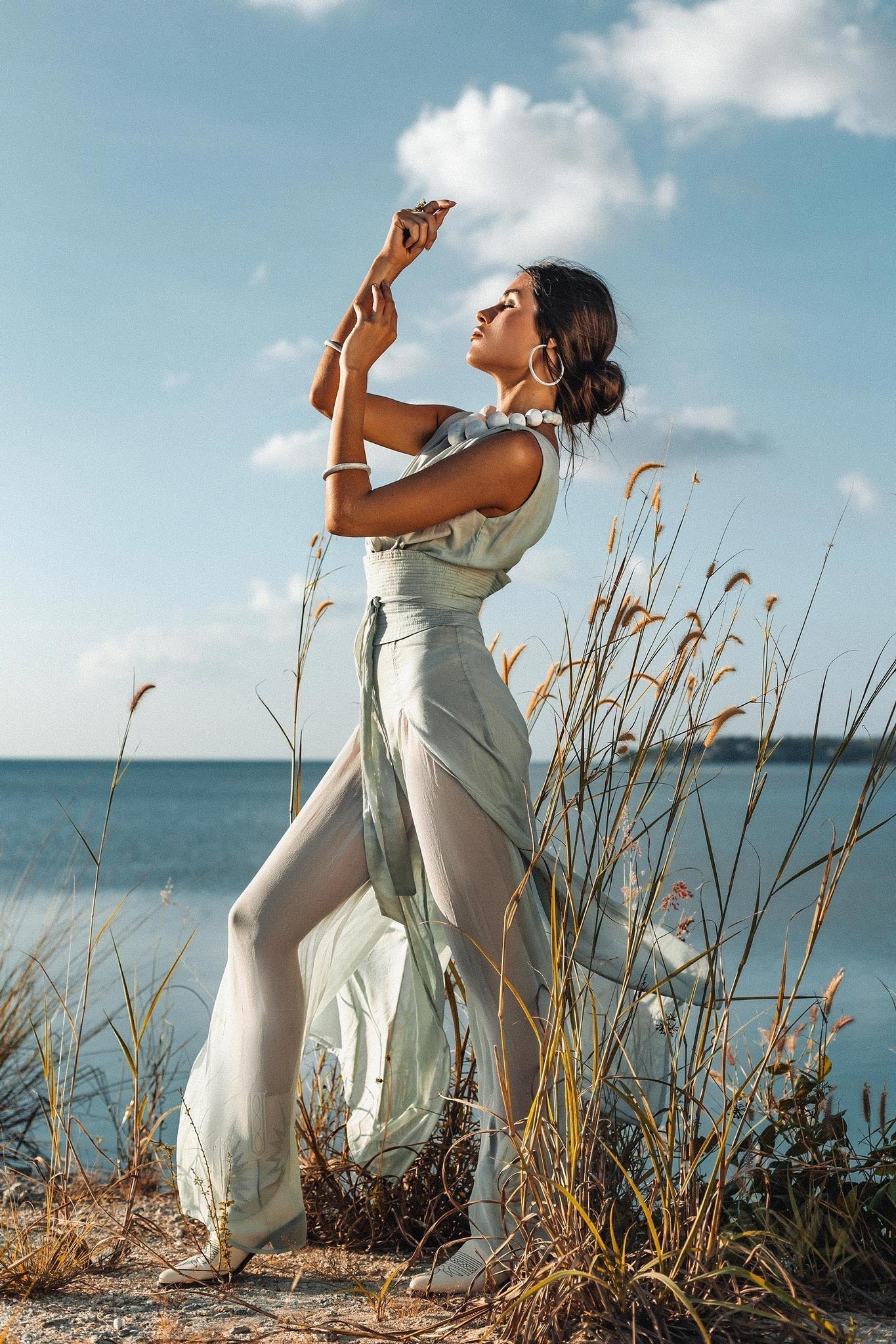 A model standing on a sandy beach with arms raised, showcasing the flowing wide legs and high side slits of the Powder Blue botanical dyed jumpsuit against a cloudy sky.