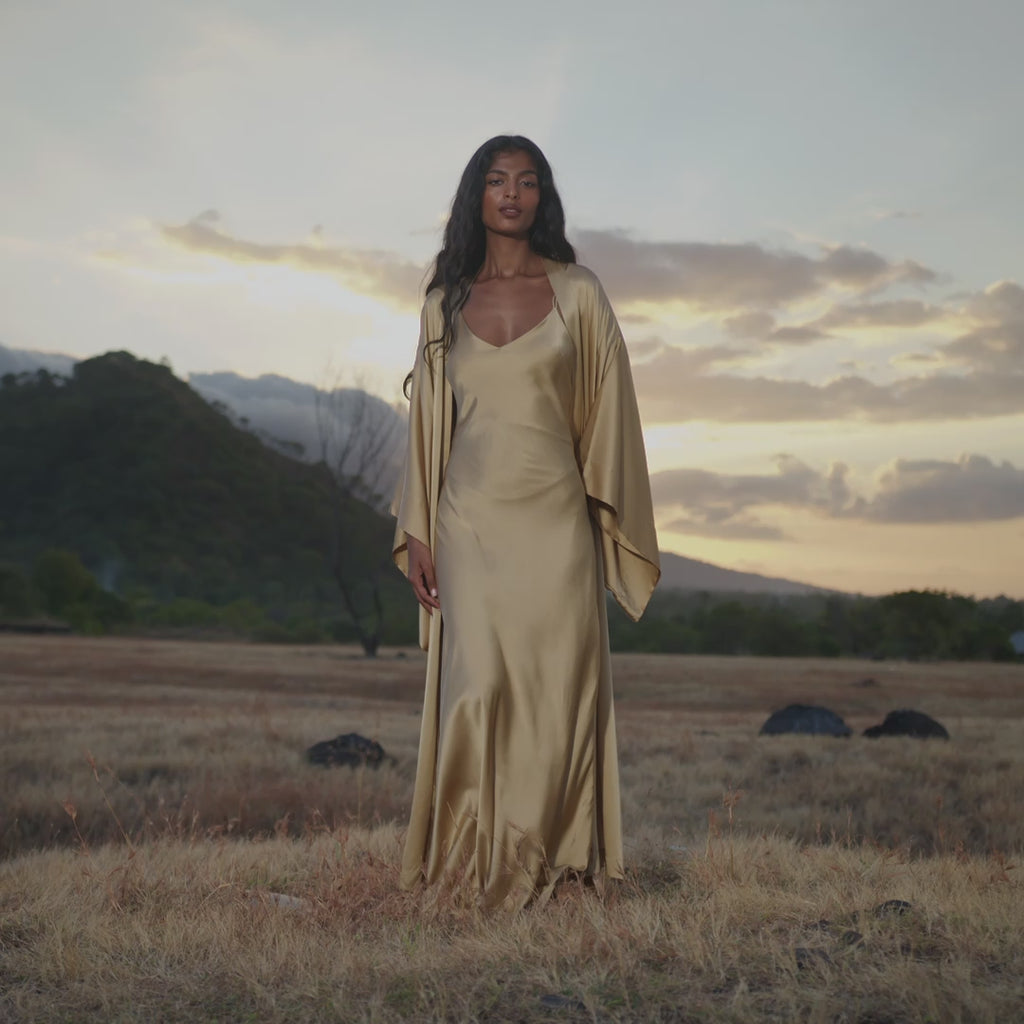 A woman in a flowing AYA Sacred Wear Gold Silk Maxi Dress with a soft V-neck and draped outer layer stands in a dry open field at sunset, with rolling hills and scattered rocks in the distance beneath a warm, clouded sky.