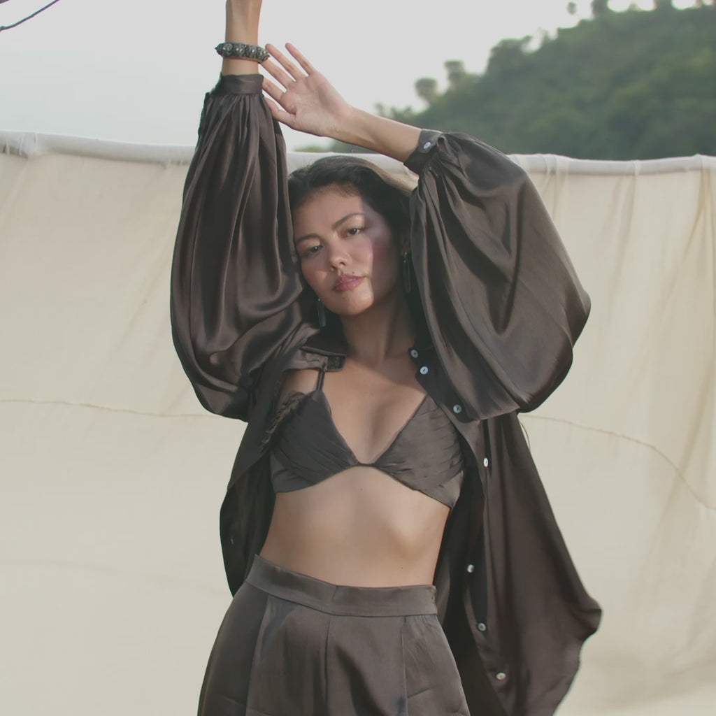 A woman poses outdoors, arms raised, in AYA Sacred Wear's Butterfly Silk Bra in Asphalt Gray — pure silk lingerie paired with matching loose layers. White fabric and a greenery hillside create a relaxed, chic atmosphere.