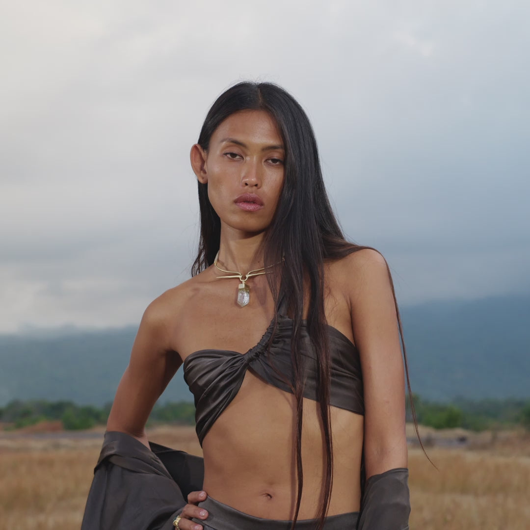 A woman stands in a field wearing a dark twisted bandeau top and Apsara Pants in Asphalt Gray – Pure Silk Flowy Pants, accessorized with rings and a necklace. Her long hair is loose, majestic mountains rising behind her.