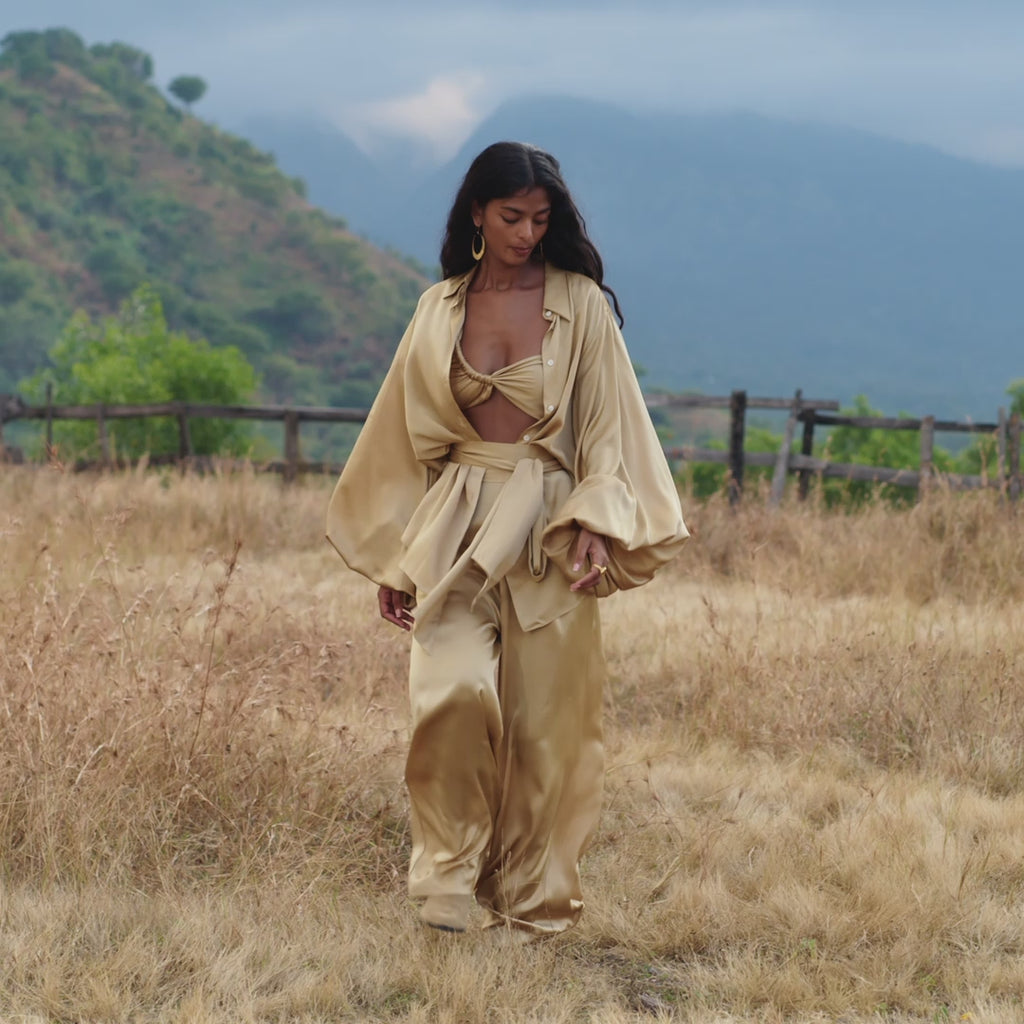 A woman with long hair wears the Apsara Kimono Blouse Inka Gold, a pure silk top by AYA Sacred Wear, standing in a dry grassy field before a wooden fence and green hills with mountains under a cloudy sky.