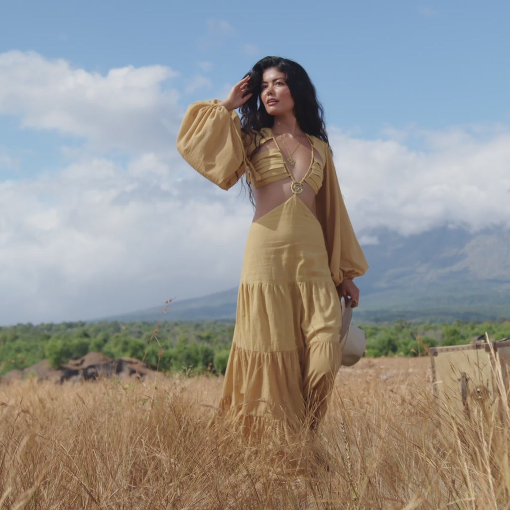 A woman smiles with eyes closed in a dry grassy field, holding a hat near a suitcase. She wears the AYA Sacred Wear Samudra Dress in Saffron Gold—an organic backless boho maxi dress. Green hills, clouds, and mountains fill the background.