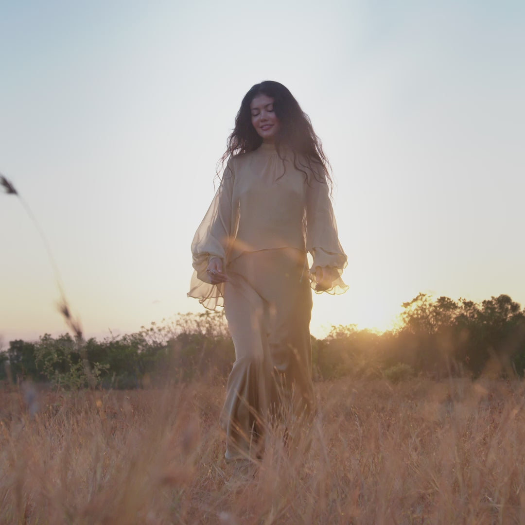 A woman with long dark hair walks through a dry, grassy field at sunset, wearing the Maharani Blouse in Inka Gold — a flowing pure silk chiffon boho top — with the sun shining behind her and trees in the background.