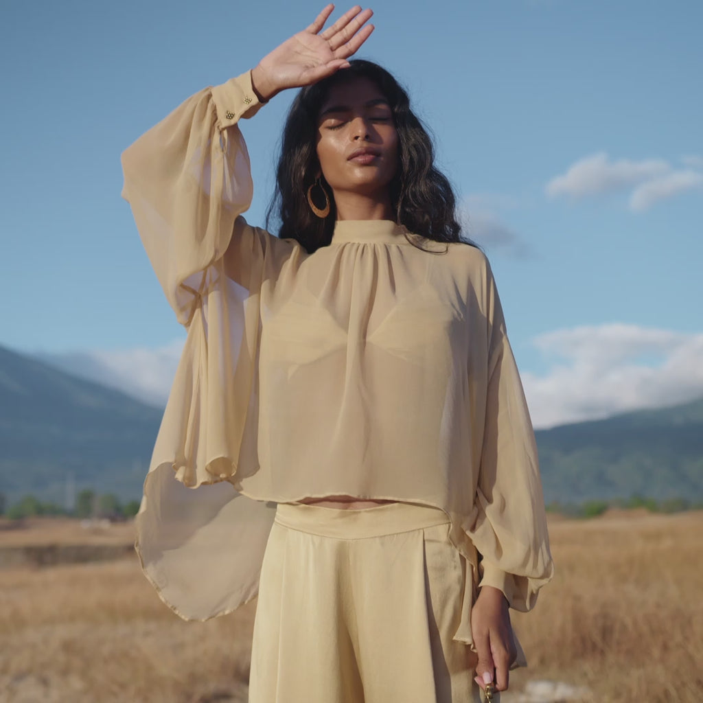 A woman in flowing Apsara Pants Inka Gold – Pure Silk Flowy Pants stands in a dry grassy field with mountains and blue sky behind her, the handcrafted Bali-inspired outfit enhancing her serene presence.