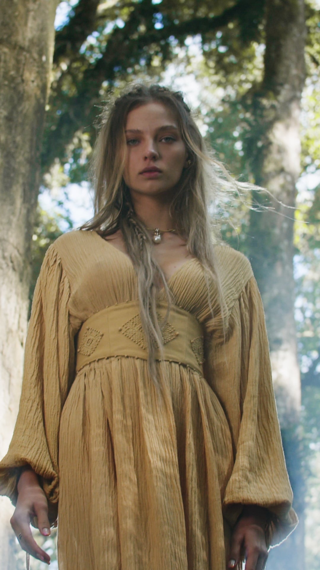 A young woman with long, wavy, light brown hair stands outdoors among trees, wearing the AYA Sacred Wear Tatva Dress in Saffron Gold – Organic Goddess Dresses. Sunlight filters through the leaves, surrounding her in a soft glow.