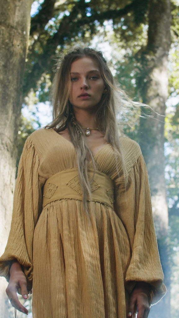 A young woman with long, wavy, light brown hair stands outdoors among trees, wearing the AYA Sacred Wear Tatva Dress in Saffron Gold – Organic Goddess Dresses. Sunlight filters through the leaves, surrounding her in a soft glow.