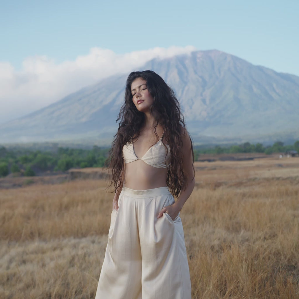 A woman with long wavy hair stands in a dry grassy field, eyes closed, wearing the AYA Sacred Wear Butterfly Silk Bra Cream – a handmade pure silk bralette. A tall mountain and green foothills create a peaceful, natural backdrop.
