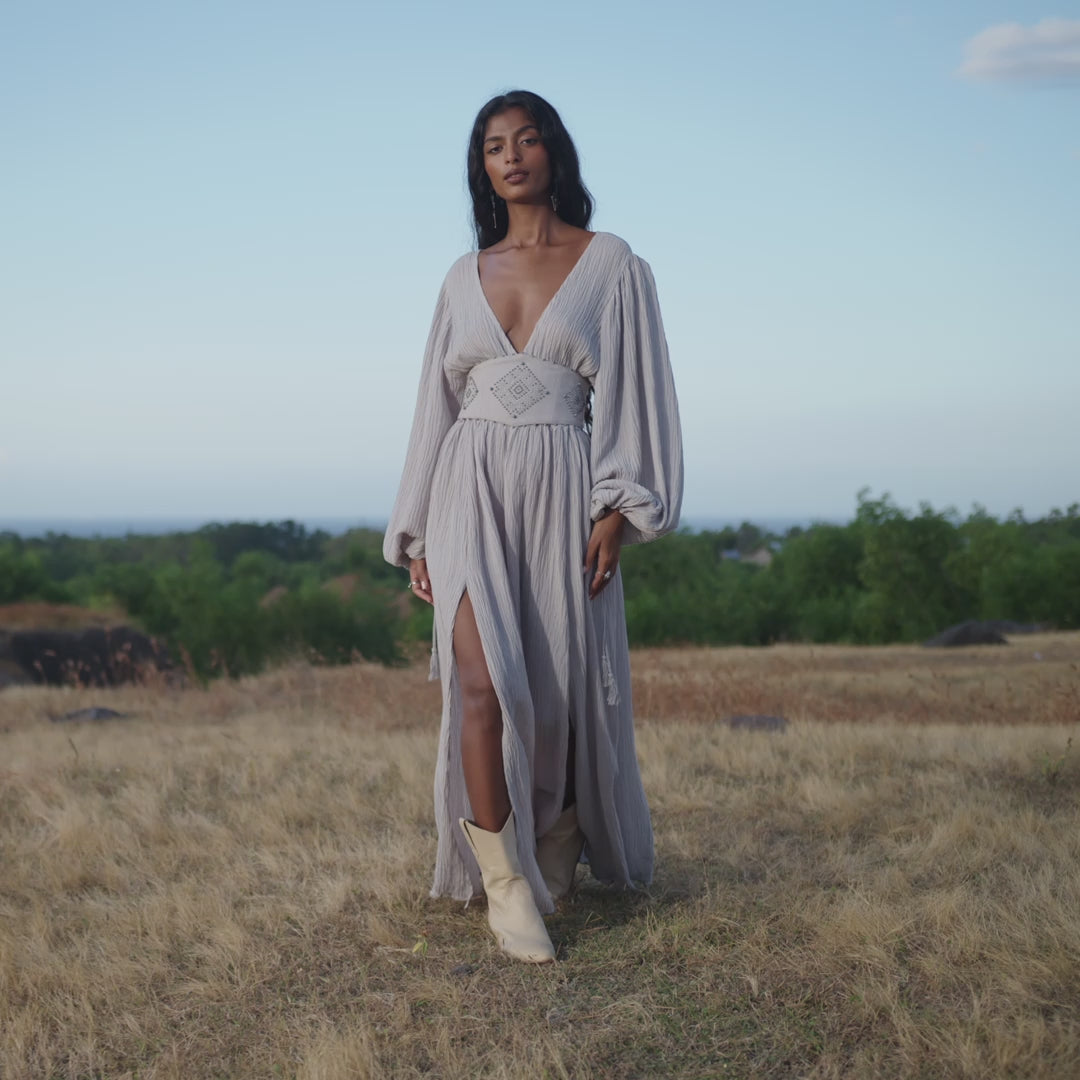 A woman with long dark hair stands on dry grass, wearing the Tatva Dress in Celestial Mist Blue by AYA Sacred Wear—a botanically dyed maxi with a deep V-neck and high slit—paired with cream boots and a handcrafted belt, framed by green trees and blue sky.