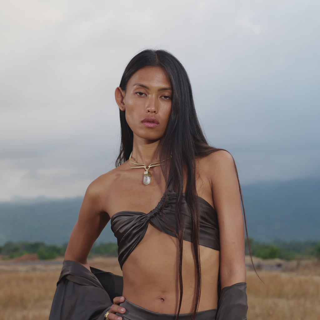 A person with long dark hair stands outdoors before mountains and a cloudy sky, wearing the AYA Sacred Wear Apsara Kimono Blouse in Asphalt Gray — an artisan pure silk kimono from Bali — draped off the shoulders, plus a twisted bandeau and crystal pendant.