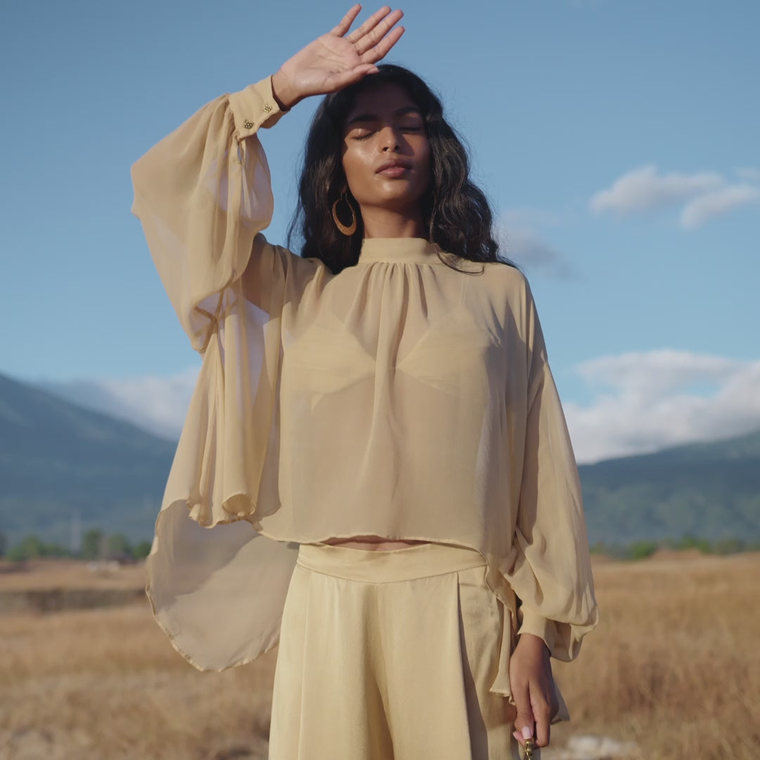 A woman stands in a sunlit field, wearing an AYA Sacred Wear Butterfly Silk Bra Inka Gold — pure silk, handmade — under a loose golden blouse and matching pants. She shades her eyes as mountains, grass, and a blue sky fill the background.