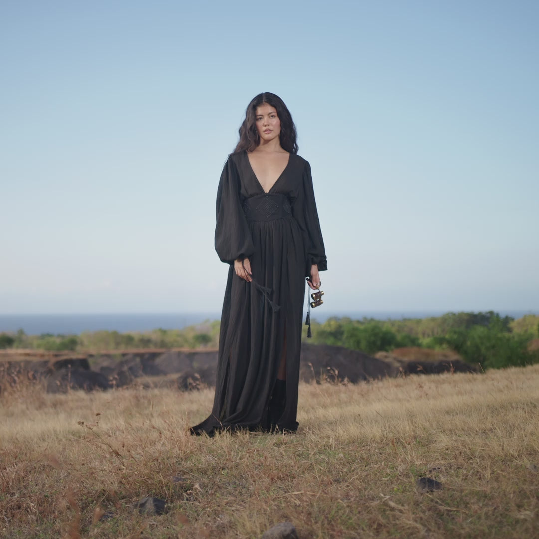 A woman stands in a dry grassy field wearing the AYA Sacred Wear Tatva Dress Black Colour – a handmade V front back kaftan with loose sleeves. She holds binocular, gazing calmly at the camera as sky and sea stretch beyond.