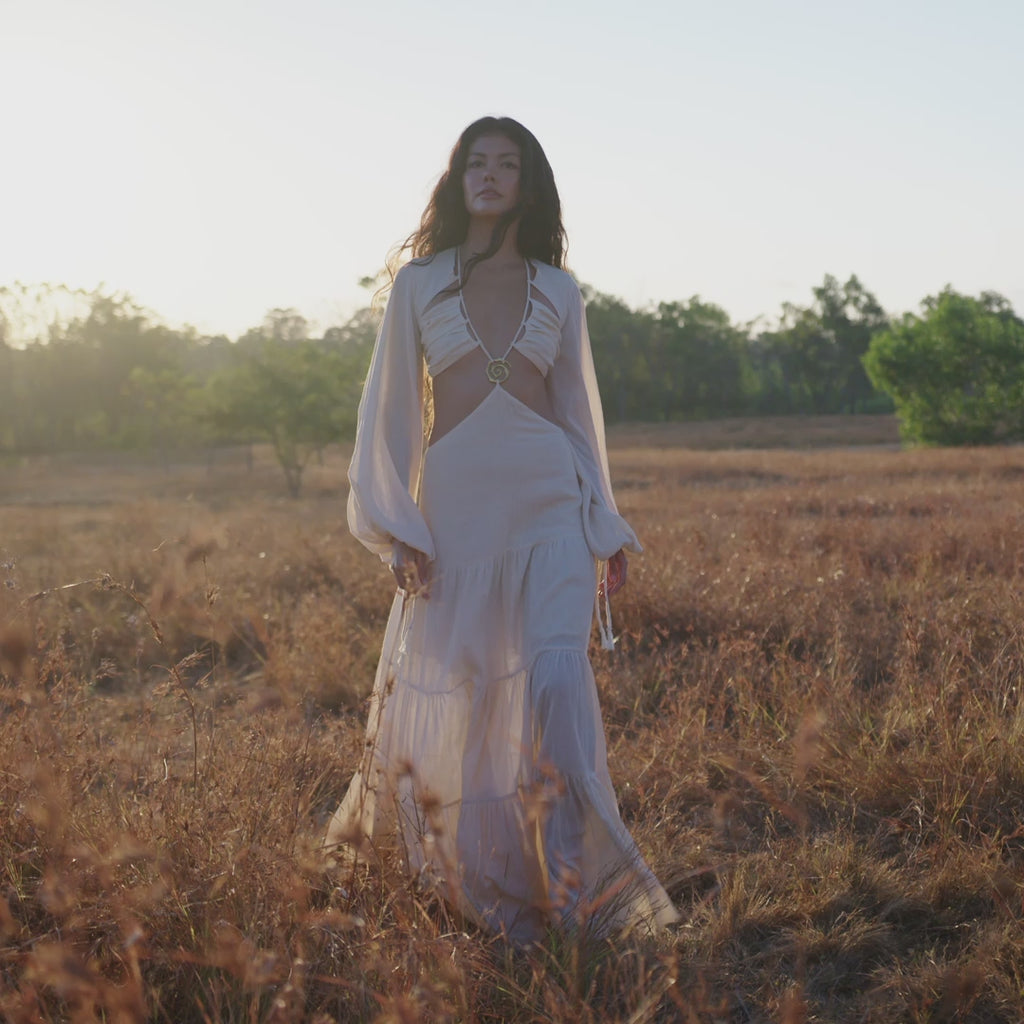 A woman with long dark hair strolls through a sunlit field at sunset in the Samudra Dress in Ivory by AYA Sacred Wear, an organic bohemian dress featuring a linen cotton blend and handmade brass spiral accents.