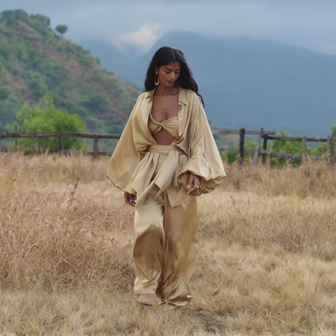 A woman with long dark hair and large hoop earrings walks thoughtfully through a grassy field wearing the Apsara Bra in Inka Gold – Pure Silk Transformable by AYA Sacred Wear. The handcrafted Bali piece flows beautifully against a backdrop of hills and clouds.