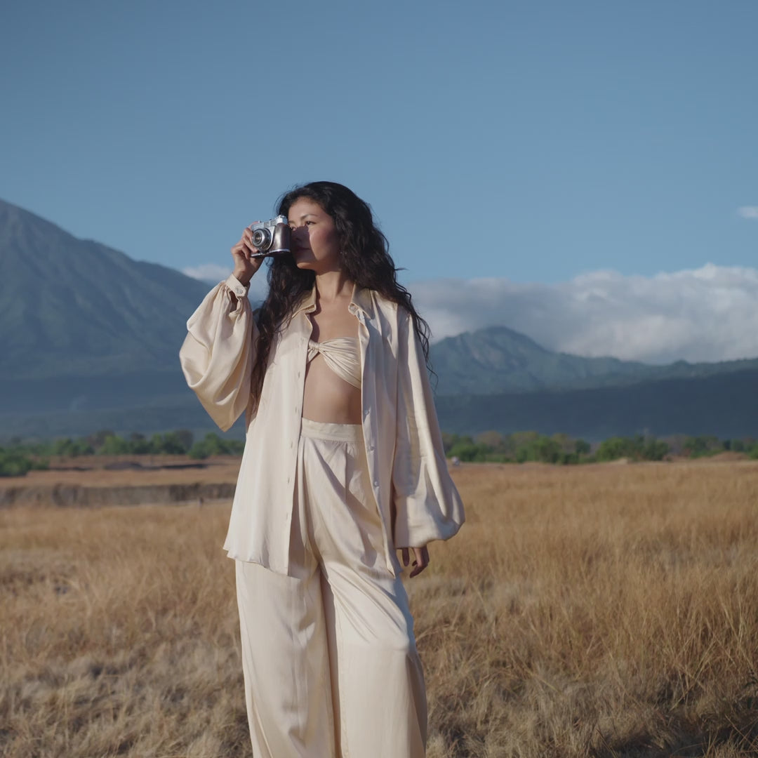 A woman in an AYA Sacred Wear Apsara Kimono Blouse Cream – Pure Silk Kimono, handcrafted in Bali, stands in a dry grassy field with a camera, long wavy hair framing her face as distant mountains and blue sky evoke natural beauty.