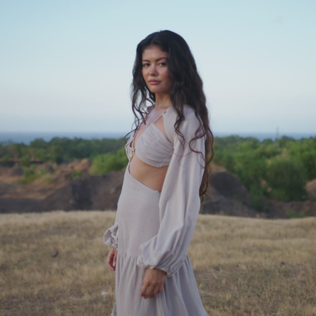 A woman with long wavy hair stands outdoors in the Samudra Dress in Celestial Mist Blue by AYA Sacred Wear, an organic bohemian garment. She gazes confidently into the distance, surrounded by greenery and a clear sky.