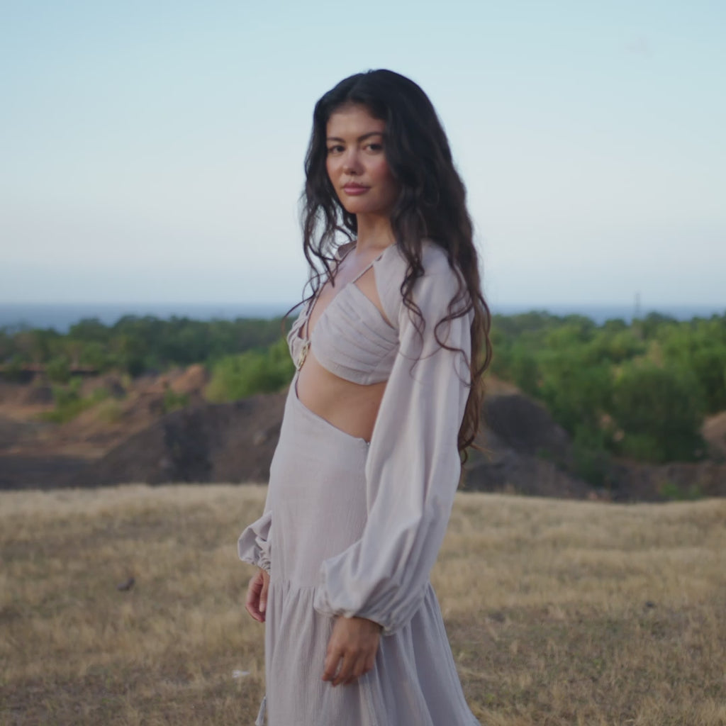 A woman with long wavy hair stands outdoors in the Samudra Dress in Celestial Mist Blue by AYA Sacred Wear, an organic bohemian garment. She gazes confidently into the distance, surrounded by greenery and a clear sky.