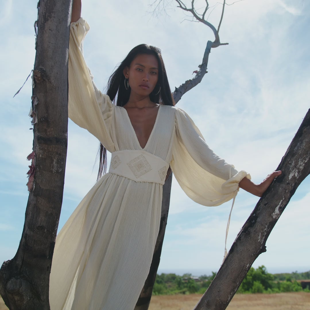 A woman with long dark hair and hoop earrings stands confidently outdoors between two bare trees, wearing the AYA Sacred Wear Tatva Dress Ivory Colour – a botanically dyed, sustainable bohemian dress paired with a handcrafted belt.