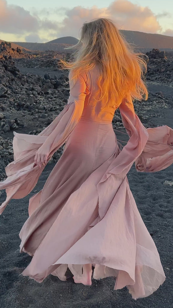 In a sunlit forest near majestic mountains, a woman stands gracefully in AYA Sacred Wear's Powder Pink Cotton Wrap Dress. The soft, organic fabric drapes elegantly, enhancing her feminine silhouette. Complemented by delicate gold jewelry, she shields her eyes with one hand, basking in the ethereal glow filtering through the lush greenery, embodying effortless bohemian elegance.