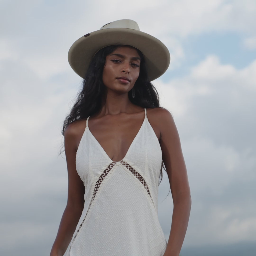 A woman stands outdoors in the AYA Sacred Wear Dhara Loom Dress in Ombre Mocco, a backless macrame pure cotton boho style with a wide-brimmed hat, posing by a log amid grassy fields and misty mountains beneath a cloudy sky.