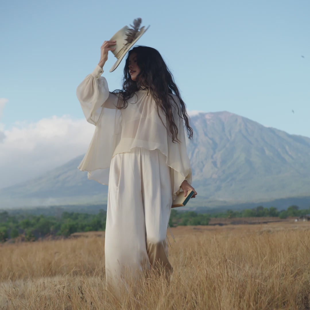 A woman with long dark hair and a wide-brimmed feathered hat stands in a dry grassy field wearing the Maharani Blouse in Cream Colour – Pure Silk Chiffon Boho Top.