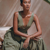 A person with short hair sits gracefully, wearing an AYA Sacred Wear Boho Bridesmaid Dress in sage green, featuring a sleeveless design and a deep V-neck with ruffled layers. They accessorize with hoop earrings and bangles, posing in gentle natural light with a blurred tropical leaf in the foreground.