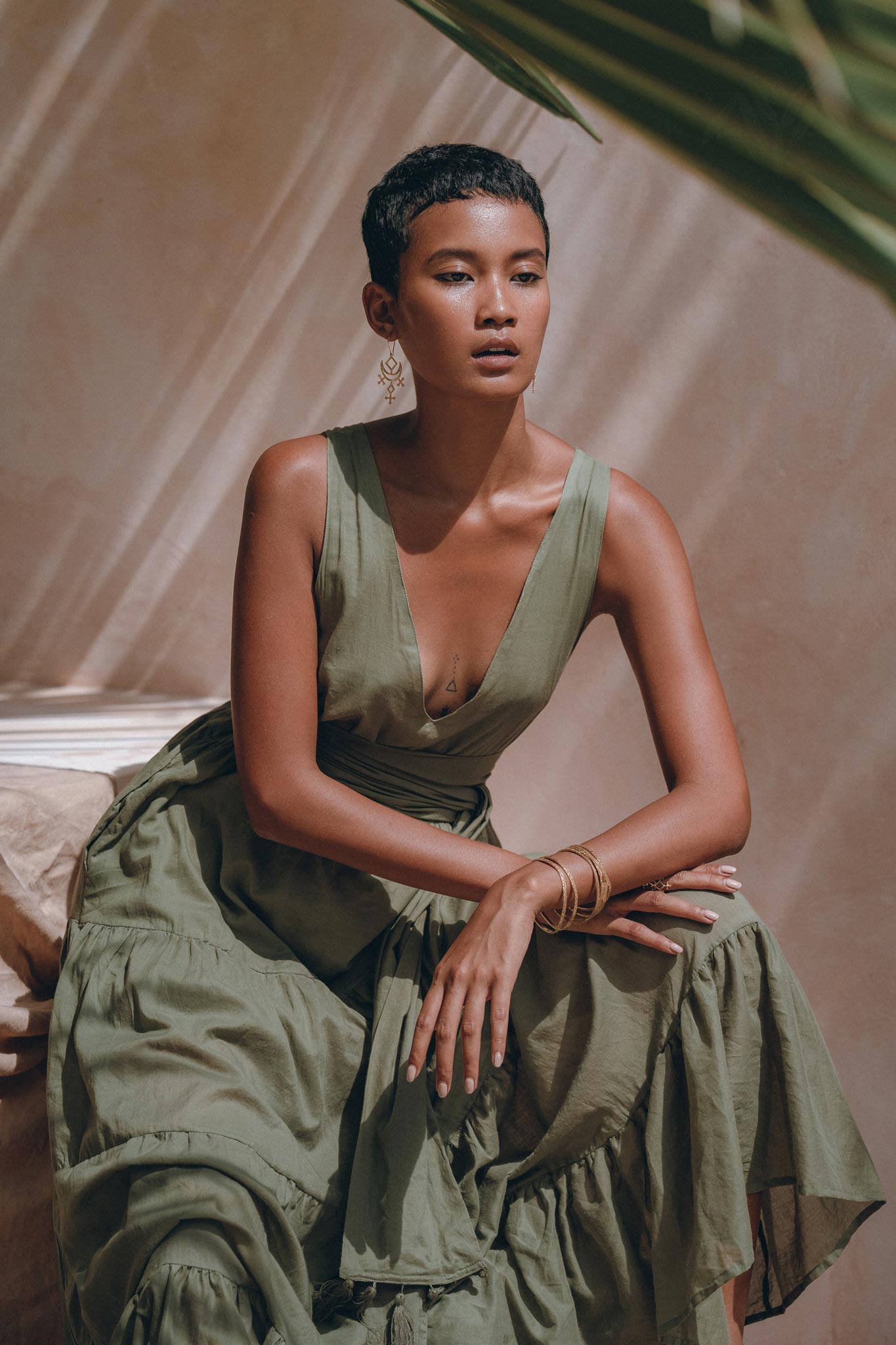 A person with short hair sits gracefully, wearing an AYA Sacred Wear Boho Bridesmaid Dress in sage green, featuring a sleeveless design and a deep V-neck with ruffled layers. They accessorize with hoop earrings and bangles, posing in gentle natural light with a blurred tropical leaf in the foreground.