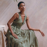 A woman in an AYA Sacred Wear Boho Bridesmaid Dress, a stylish Sage Green Adjustable Bohemian Dress, sits elegantly on a textured surface. She accessorizes with gold hoop earrings and bracelets, set against the backdrop of softly hued earth-toned walls and a green leaf overhead, creating a serene atmosphere.