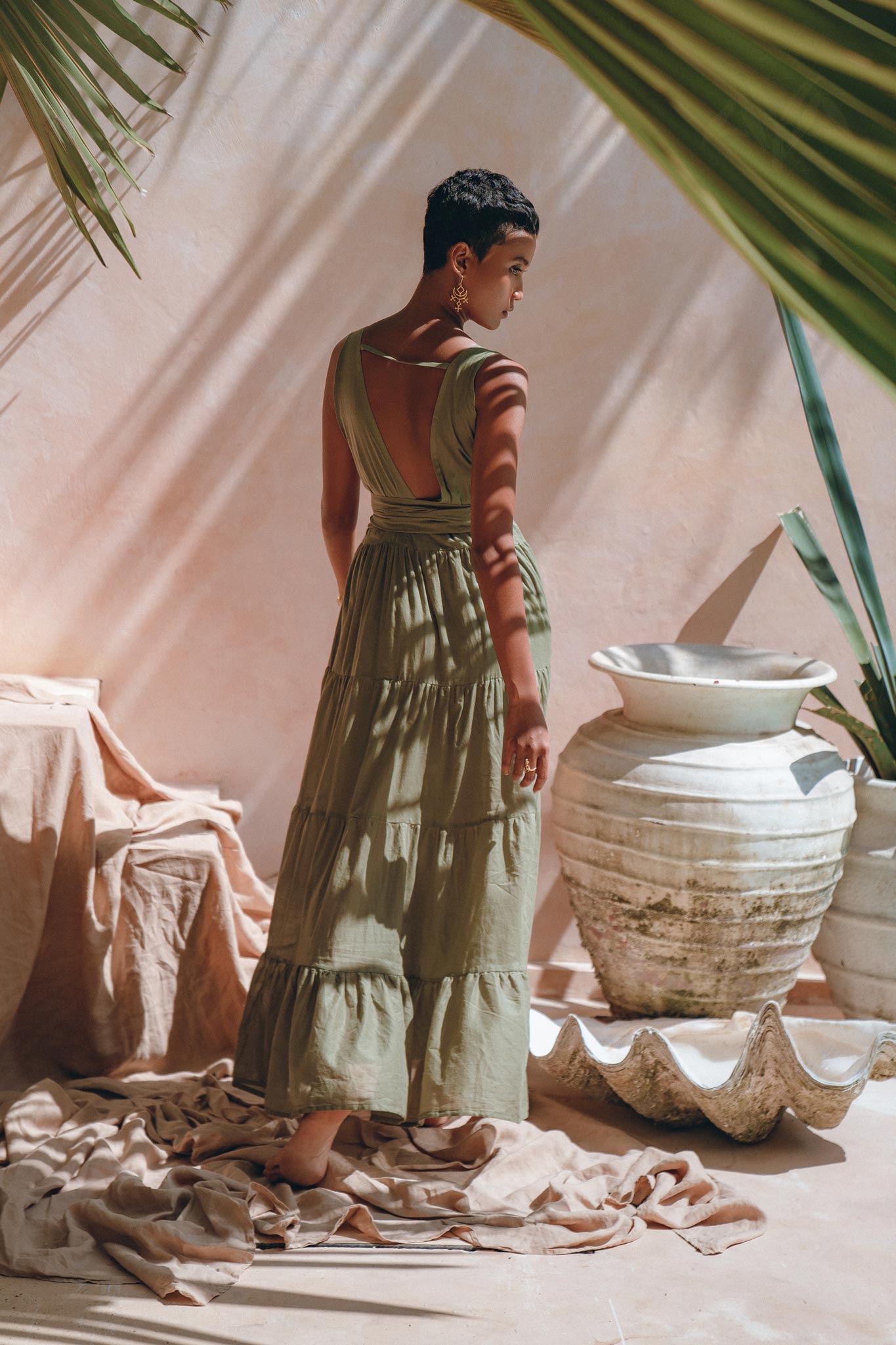 A person stands with their back to the camera, wearing a sage green Boho Bridesmaid Dress by AYA Sacred Wear. They are in a sunlit space adorned with large ceramic vases, beige fabric, and tropical leaves casting shadows around them.