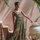 In a sunlit room with soft shadows, an individual wearing a long, flowing Sage Green Adjustable Dress by AYA Sacred Wear stands gracefully. They are positioned beside a large decorative vase while palm leaves hang overhead, enhancing the serene and elegant ambiance.
