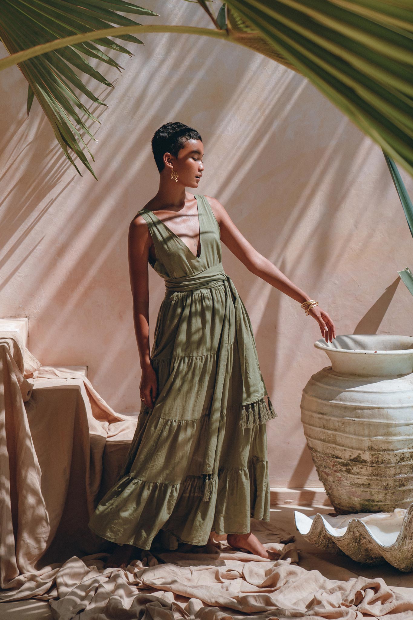 In a sunlit room with soft shadows, an individual wearing a long, flowing Sage Green Adjustable Dress by AYA Sacred Wear stands gracefully. They are positioned beside a large decorative vase while palm leaves hang overhead, enhancing the serene and elegant ambiance.
