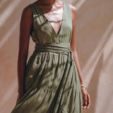 In soft lighting, a person is dressed in a sleeveless sage green Boho Bridesmaid Dress by AYA Sacred Wear. Crafted from organic cotton, the dress showcases a deep V-neck and tiered skirt, with an adjustable sash cinching the waist. The individual sports short hair adorned with earrings and bracelets, casting shadows on a light background.