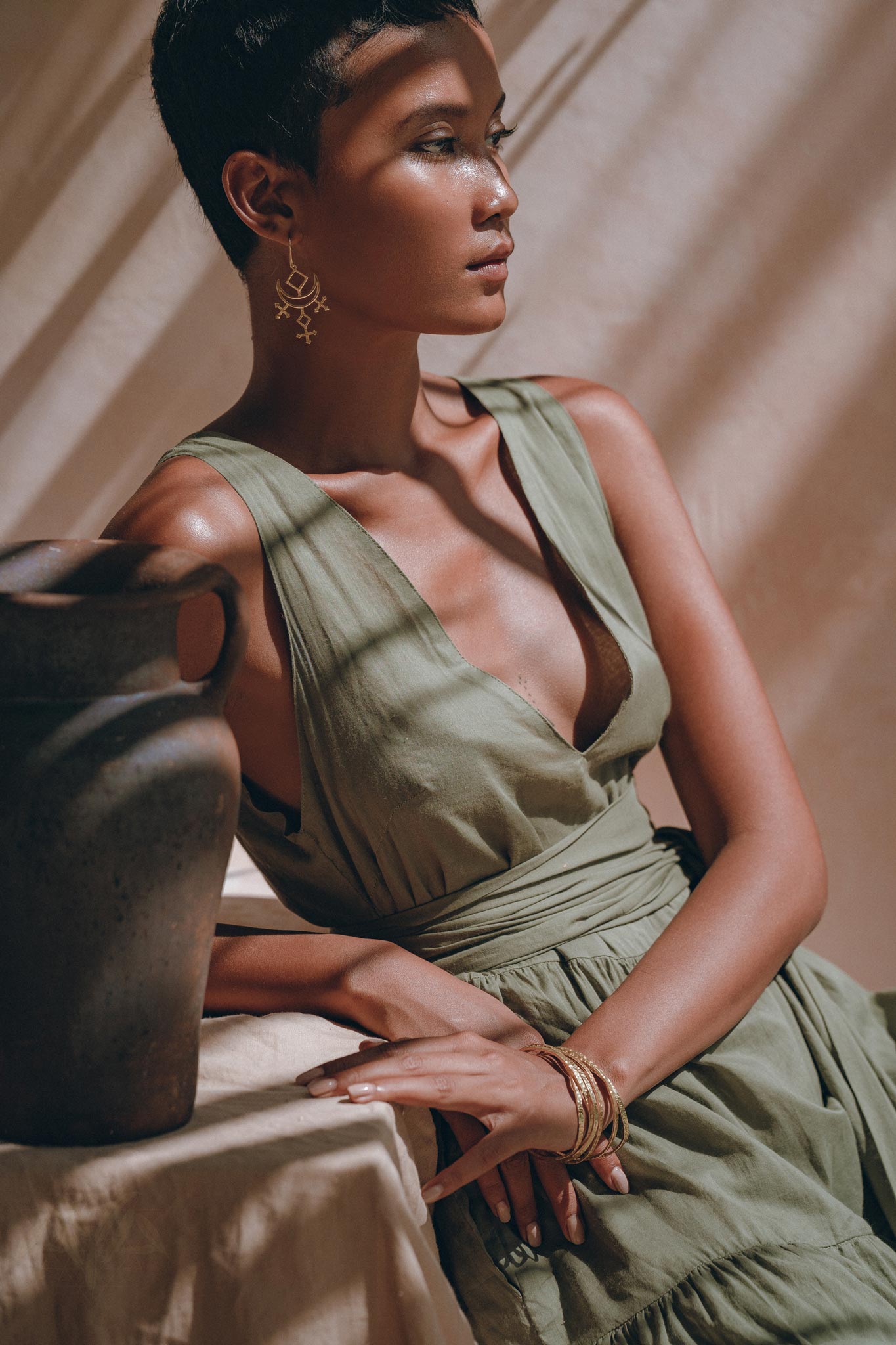 A woman in an AYA Sacred Wear Boho Bridesmaid Dress, a Sage Green Adjustable Bohemian Dress, sits beside a clay pot on a table. She has short hair and wears large earrings along with bracelets. Sunlight casts shadows across her and her surroundings, creating a dappled effect.